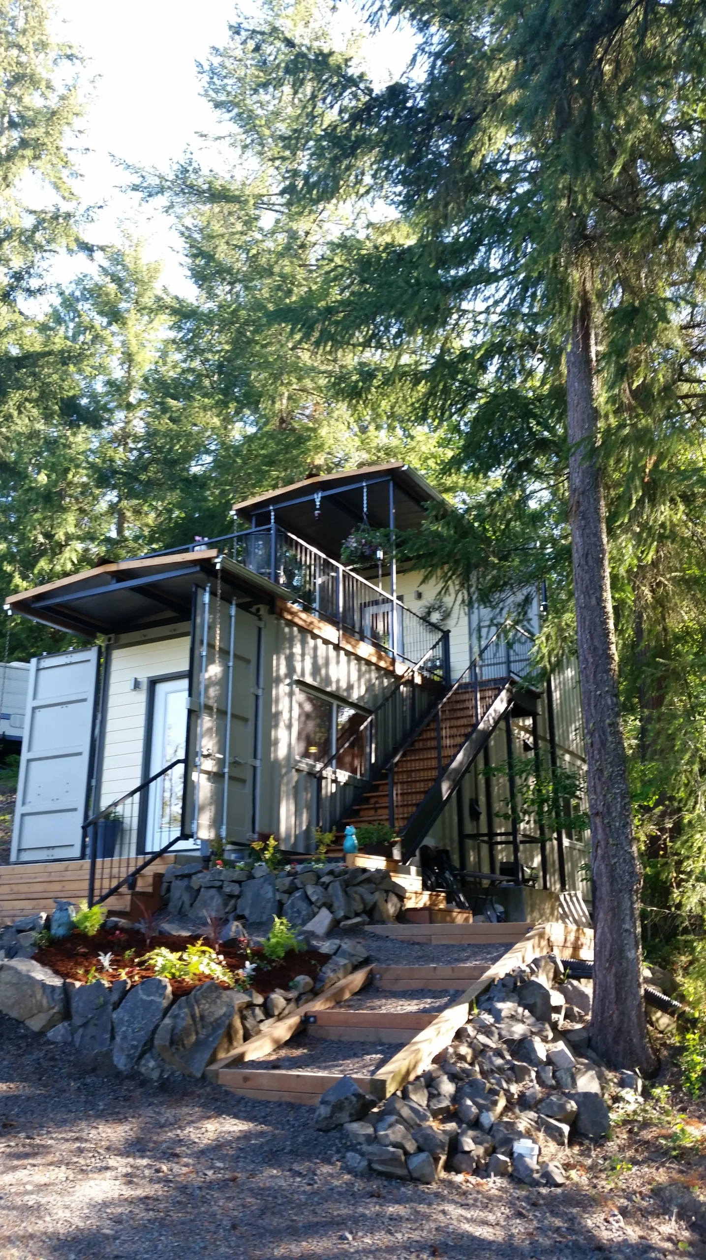 Highland & Co. Acres completed shipping container tiny home nestled in forest with multi-level decks, wooden stairs, and natural stone landscaping
