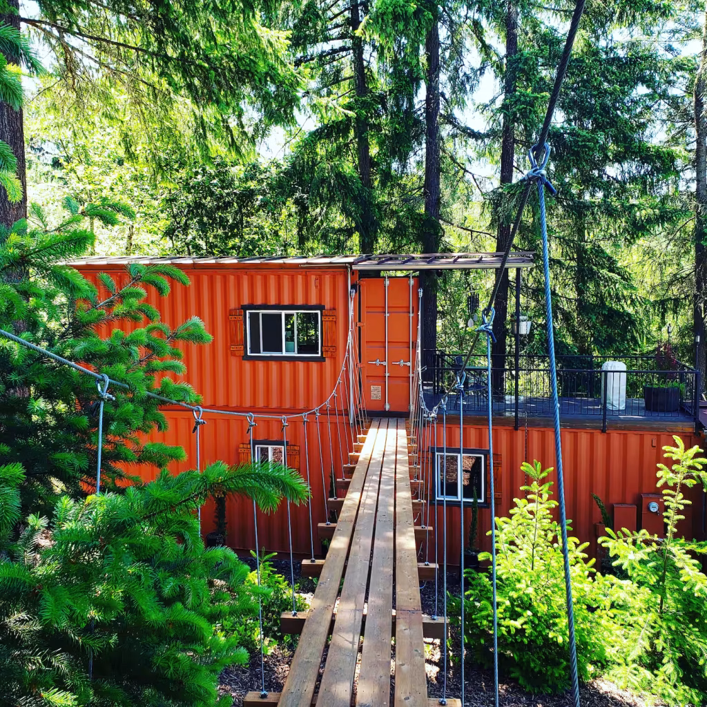 Suspension bridge leading to orange shipping container tiny home nestled in forest at Highland & Co. Acres unique Airbnb accommodation