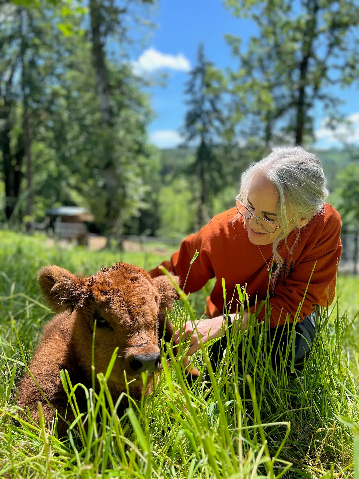 Jaimie Hinckle kneeling in tall green grass gently petting a small red Highland calf at Highland & Co. Acres farm in Cowlitz County, Washington